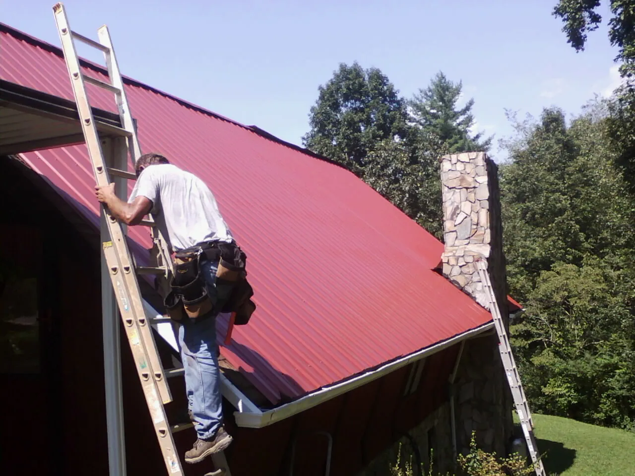 Roofer on a ladder beside a red metal roof and stone chimney — Soffit & Fascia Repair by Roofing Brothers in Huntingdon Valley